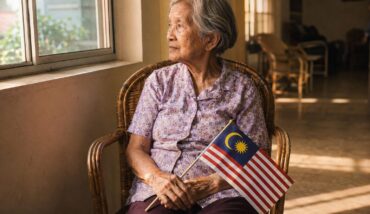 An elderly Malaysian woman sits by a window holding a small Jalur Gemilang, late afternoon light across the room.