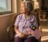 An elderly Malaysian woman sits by a window holding a small Jalur Gemilang, late afternoon light across the room.