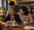 A woman laughs with her elderly mother and teenage son around a kitchen table at home